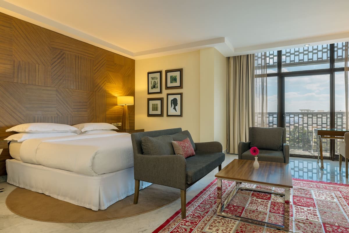 A hotel room with a bed, a sofa, a coffee table, and a view of the city through large windows.