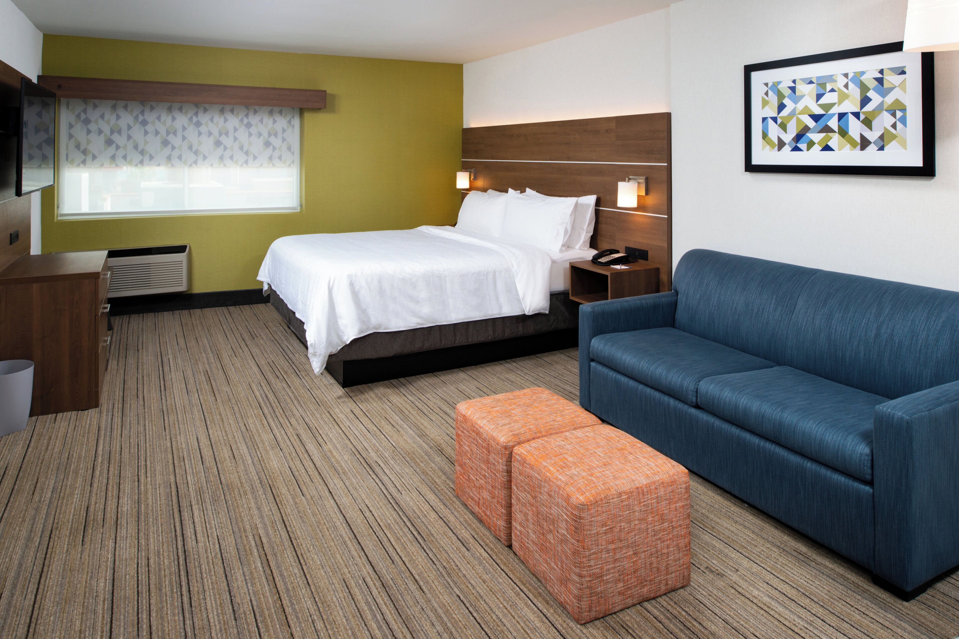 A hotel room with a bed, a blue sofa, an orange ottoman, a bedside table with a lamp, and a framed picture on the wall.