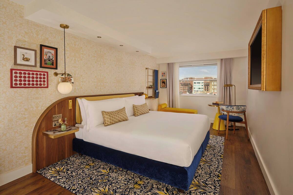 Ein Hotelzimmer mit einem großen Bett, einem Nachttisch, einem Sessel, einem Fenster mit Blick auf Gebäude und einer Wand mit gerahmten Bildern.