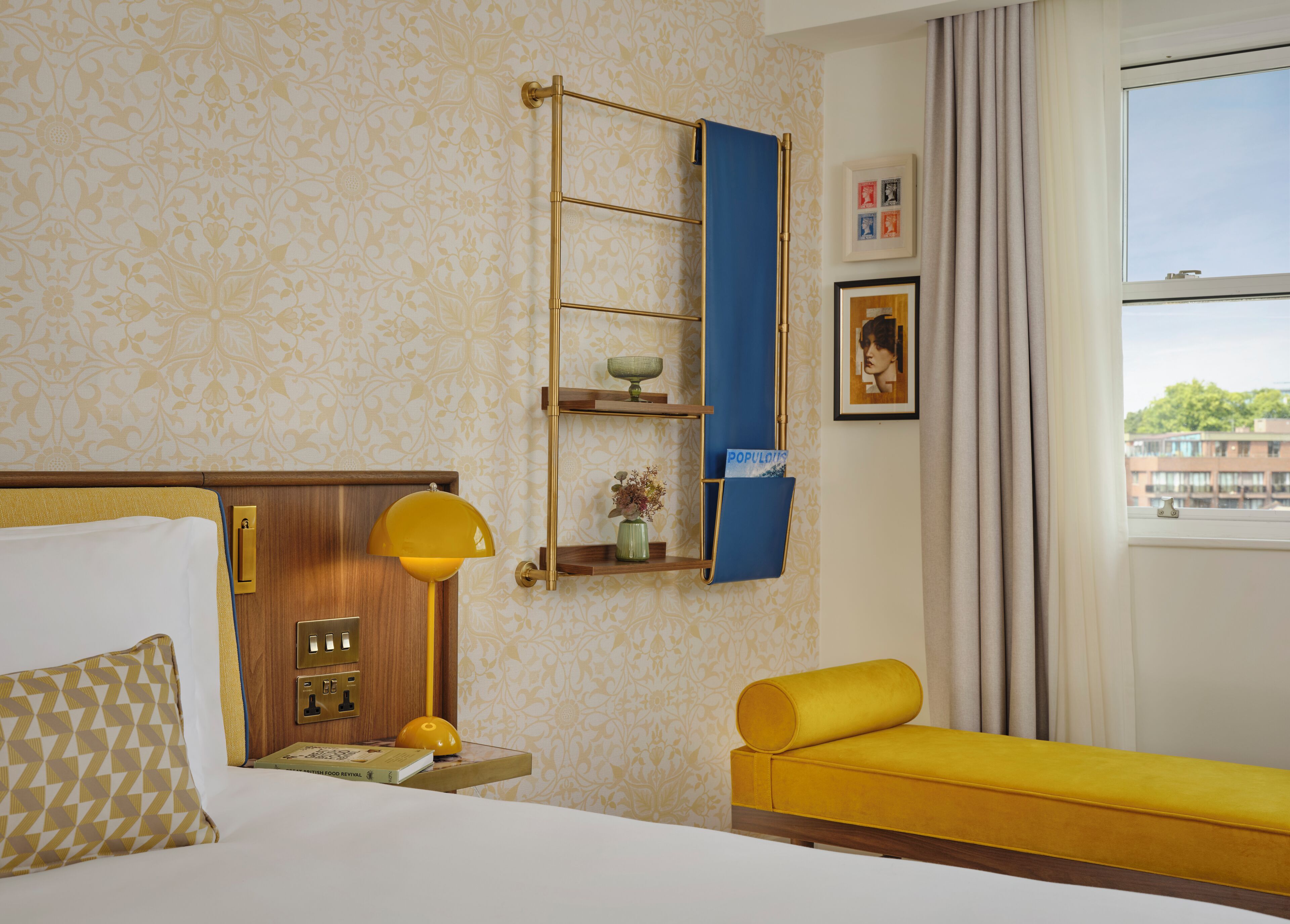 A hotel room with a bed, a bedside table with a lamp, a shelf with decorative items, a window with a view, and a yellow sofa.