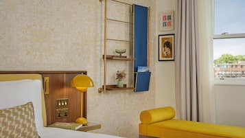 A hotel room with a bed, a bedside table with a lamp, a shelf with decorative items, a window with a view, and a yellow sofa.