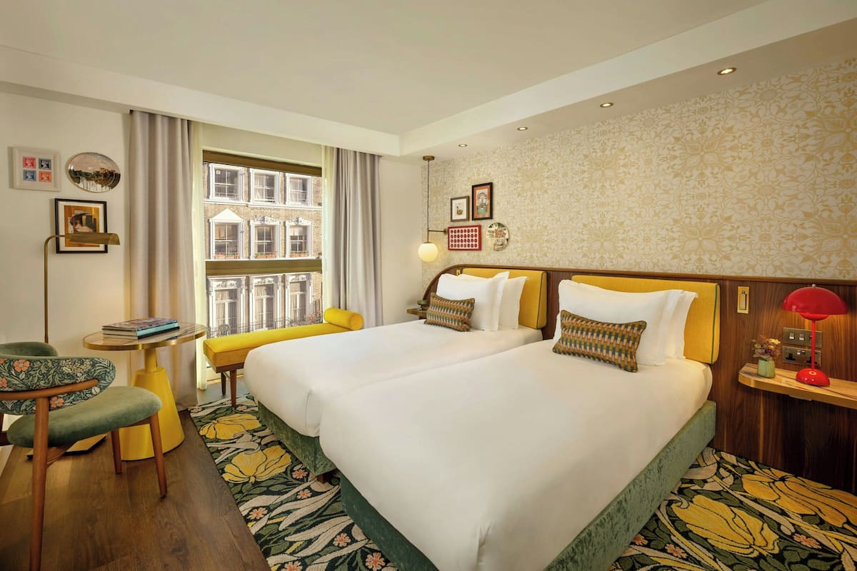 Ein Hotelzimmer mit zwei Betten, einem Schreibtisch, einem Sessel, einem Fenster mit Vorhängen und einem gemusterten Teppich.