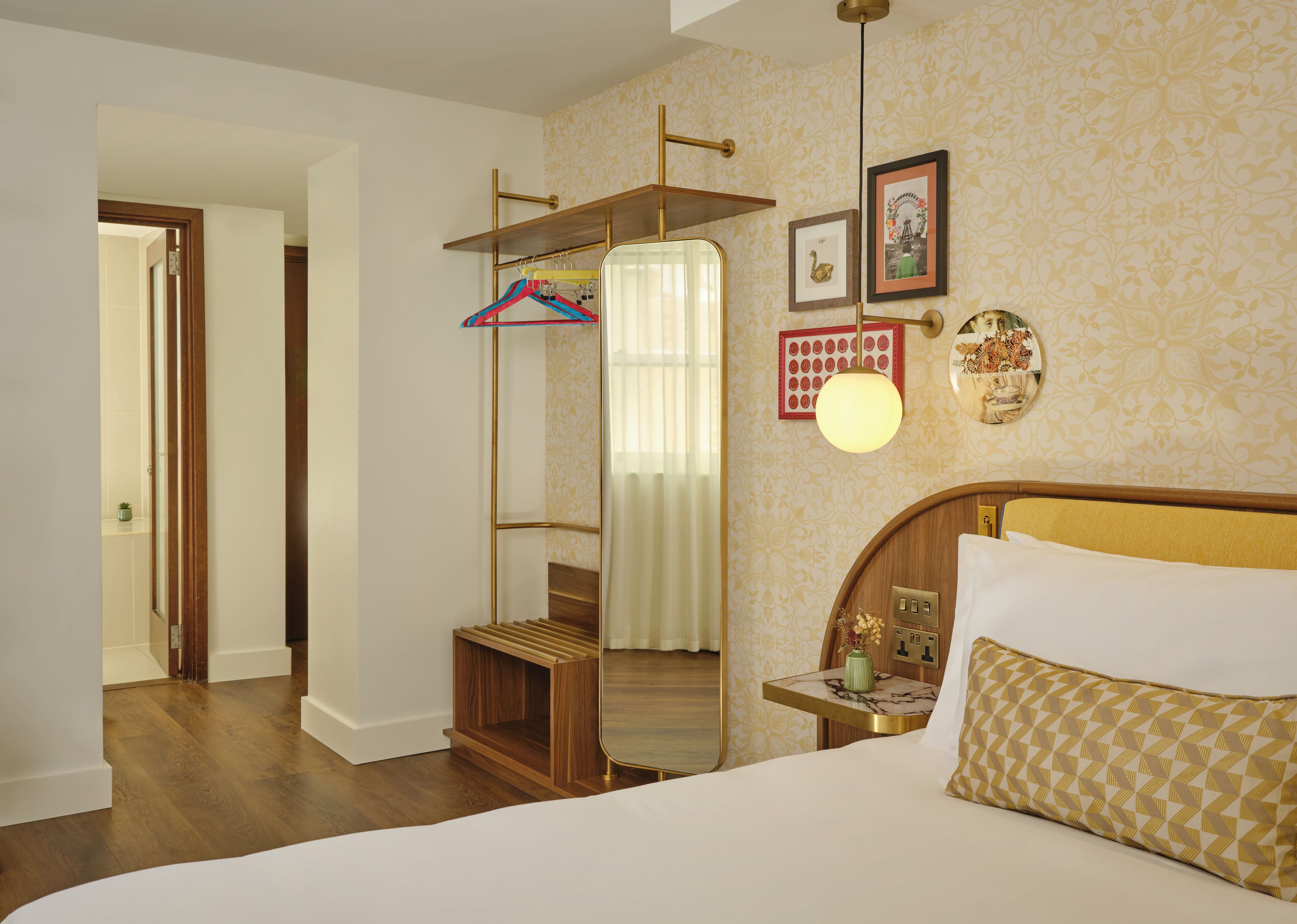 Quarto de hotel com uma cama, cabeceira de madeira, mesa de cabeceira com abajur, espelho e prateleira na parede com cabides.