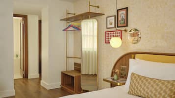Quarto de hotel com uma cama, cabeceira de madeira, mesa de cabeceira com abajur, espelho e prateleira na parede com cabides.