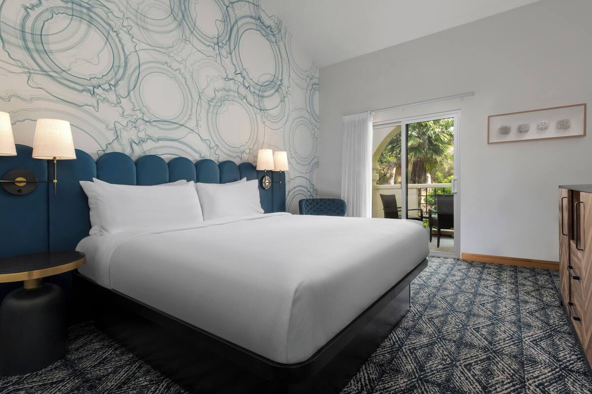 A modern hotel room with a large bed, two bedside lamps, a patterned carpet, and a view of the outdoors through a sliding door.