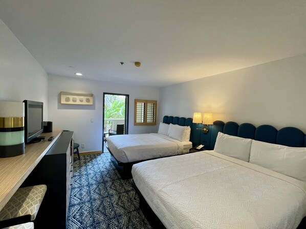 A hotel room with two beds, a desk with a television, and a view of greenery through a door.
