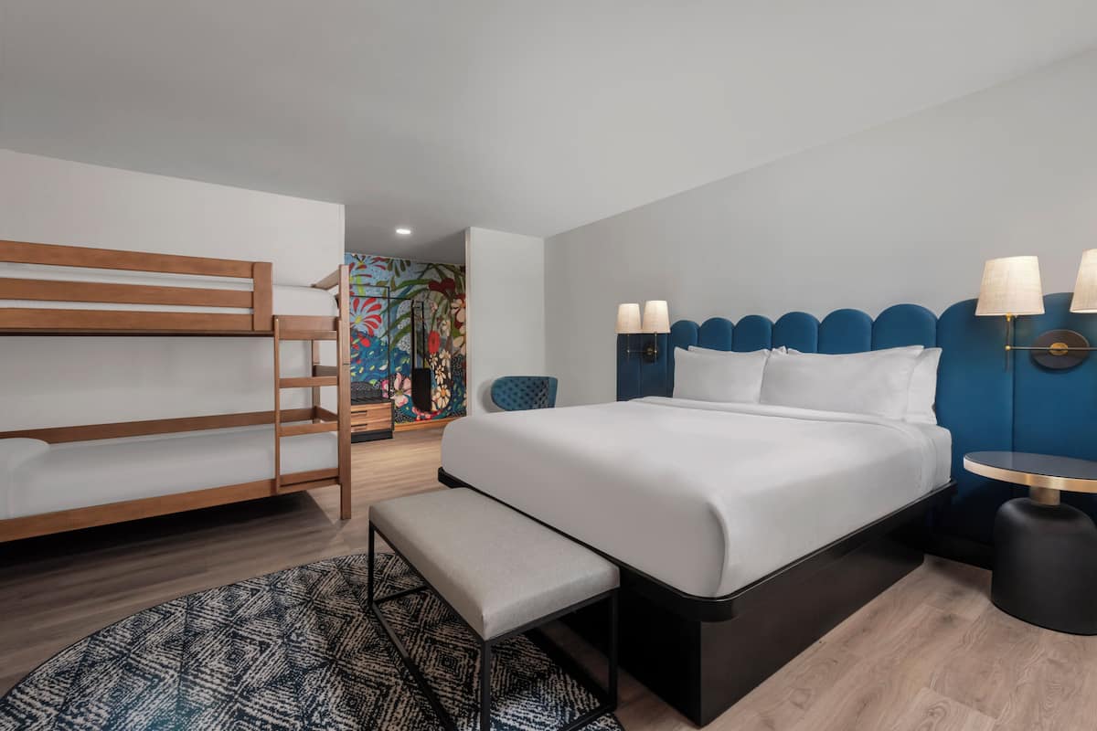 A modern hotel room with a bunk bed, a large bed with a blue headboard, a bedside table with lamps, and a patterned rug.