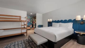 A modern hotel room with a bunk bed, a large bed with a blue headboard, a bedside table with lamps, and a patterned rug.