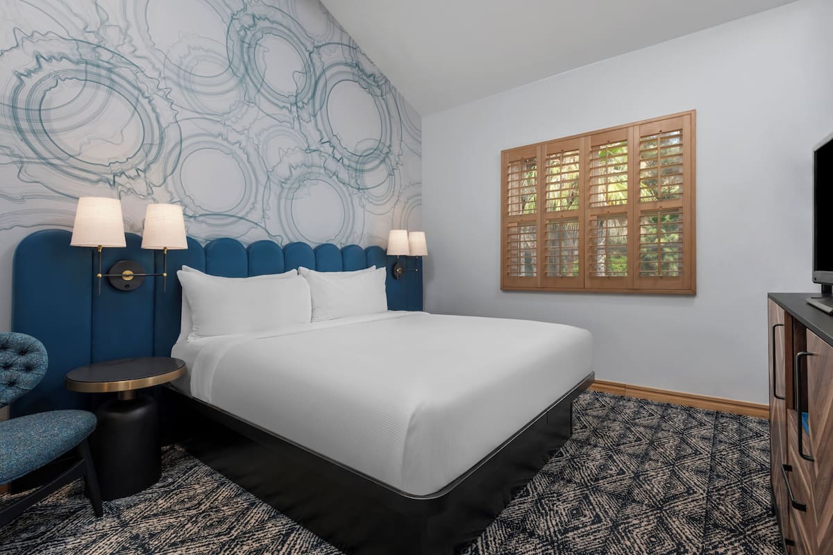 A modern bedroom with a large bed, two bedside tables with lamps, a blue headboard, a patterned rug, a wooden cabinet, and a window with a view of greenery.