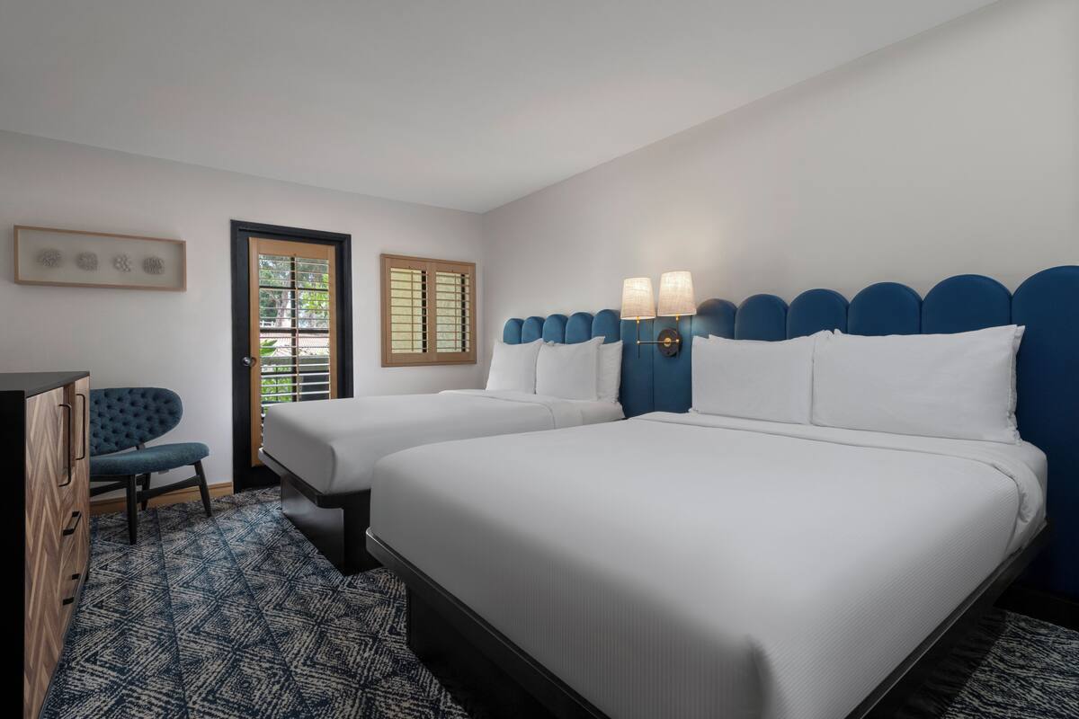 A hotel room with two beds, a desk, a chair, and a window with blinds.