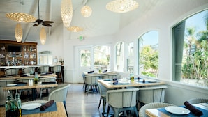 A restaurant with high ceilings, wicker furniture, and large windows offering views of greenery. - Catalina Canyon Inn (Avalon)