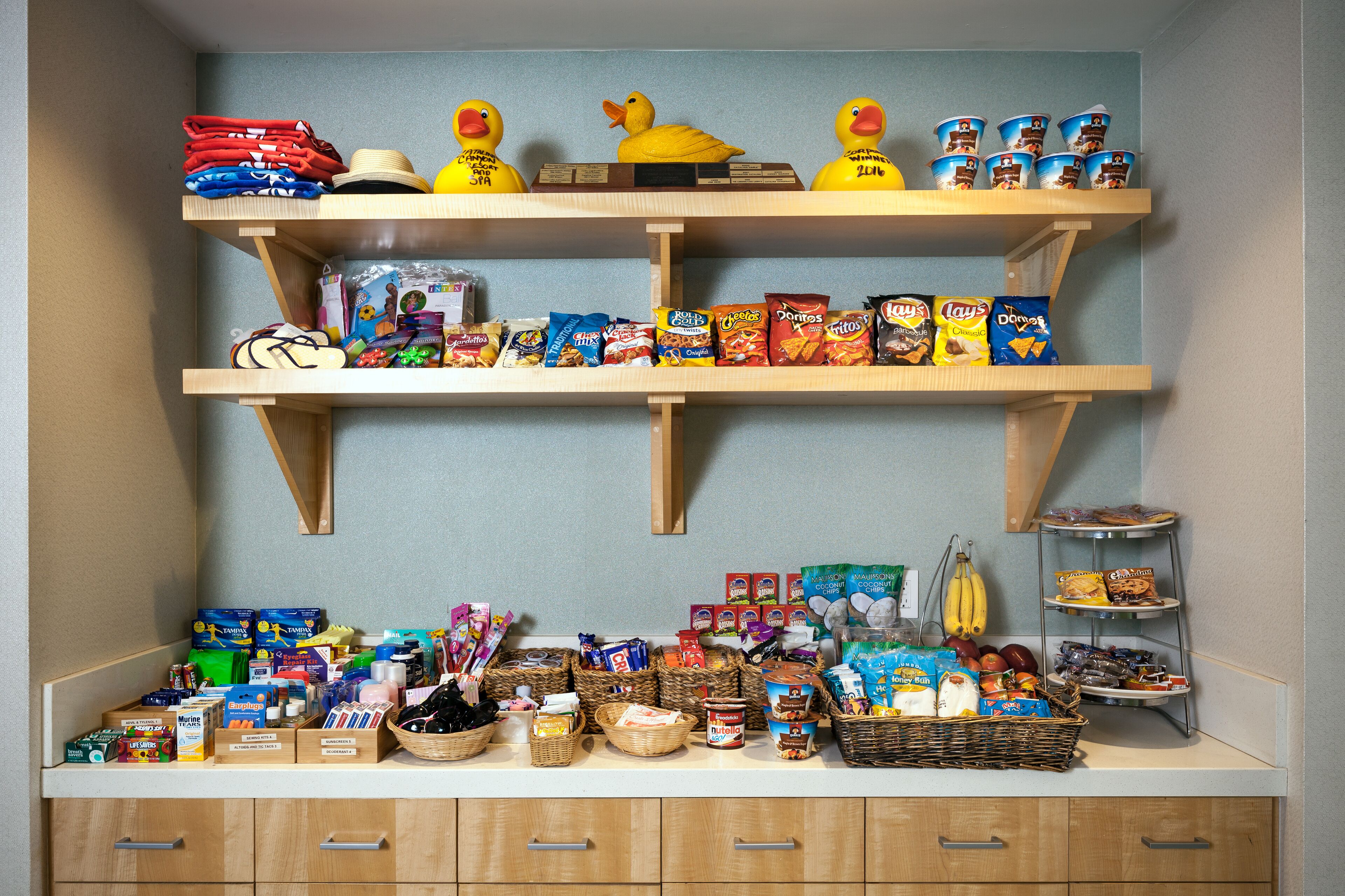 Una encimera de cocina con diversos alimentos y utensilios, incluyendo una cesta de plátanos, un tazón de cereales y una repisa con una figurita de pato amarillo.