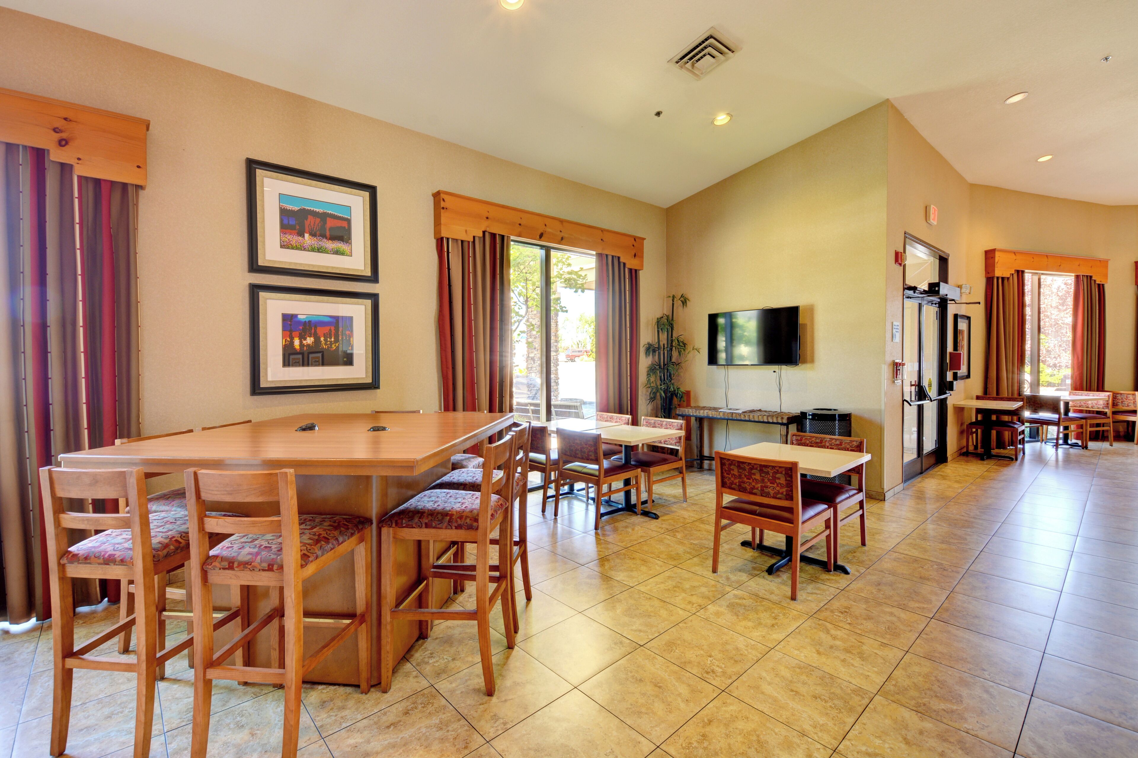 A spacious dining area with a long wooden table and multiple chairs, a television mounted on the wall, and a kitchen area with a refrigerator and stove.