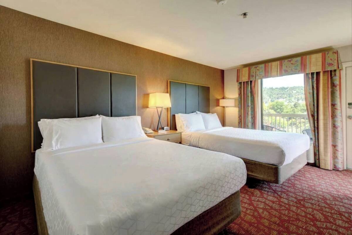 A hotel room with two beds, a large window with curtains, and a view of greenery outside.