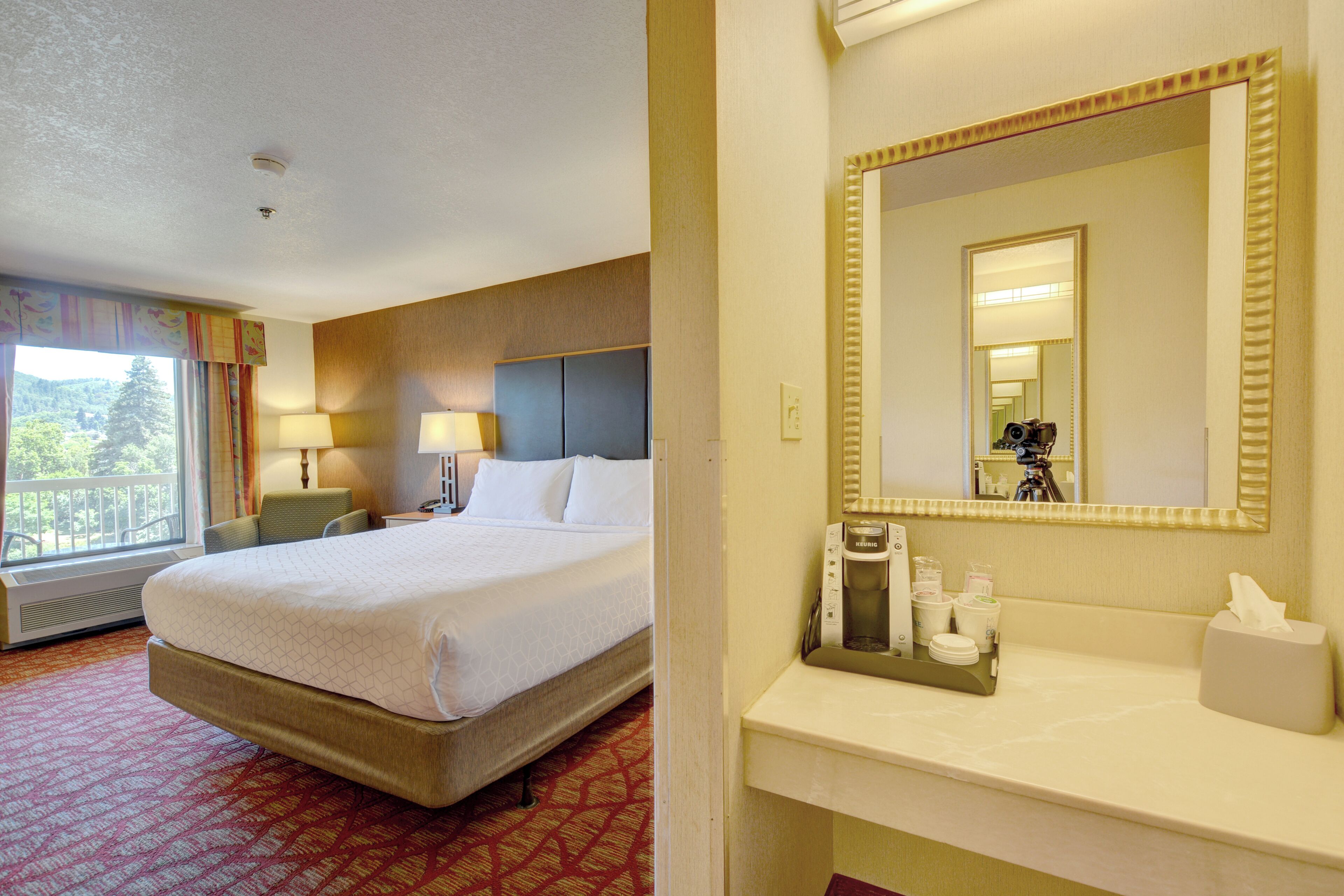 A hotel room with a large bed, a bathroom with a mirror, and a window with curtains.