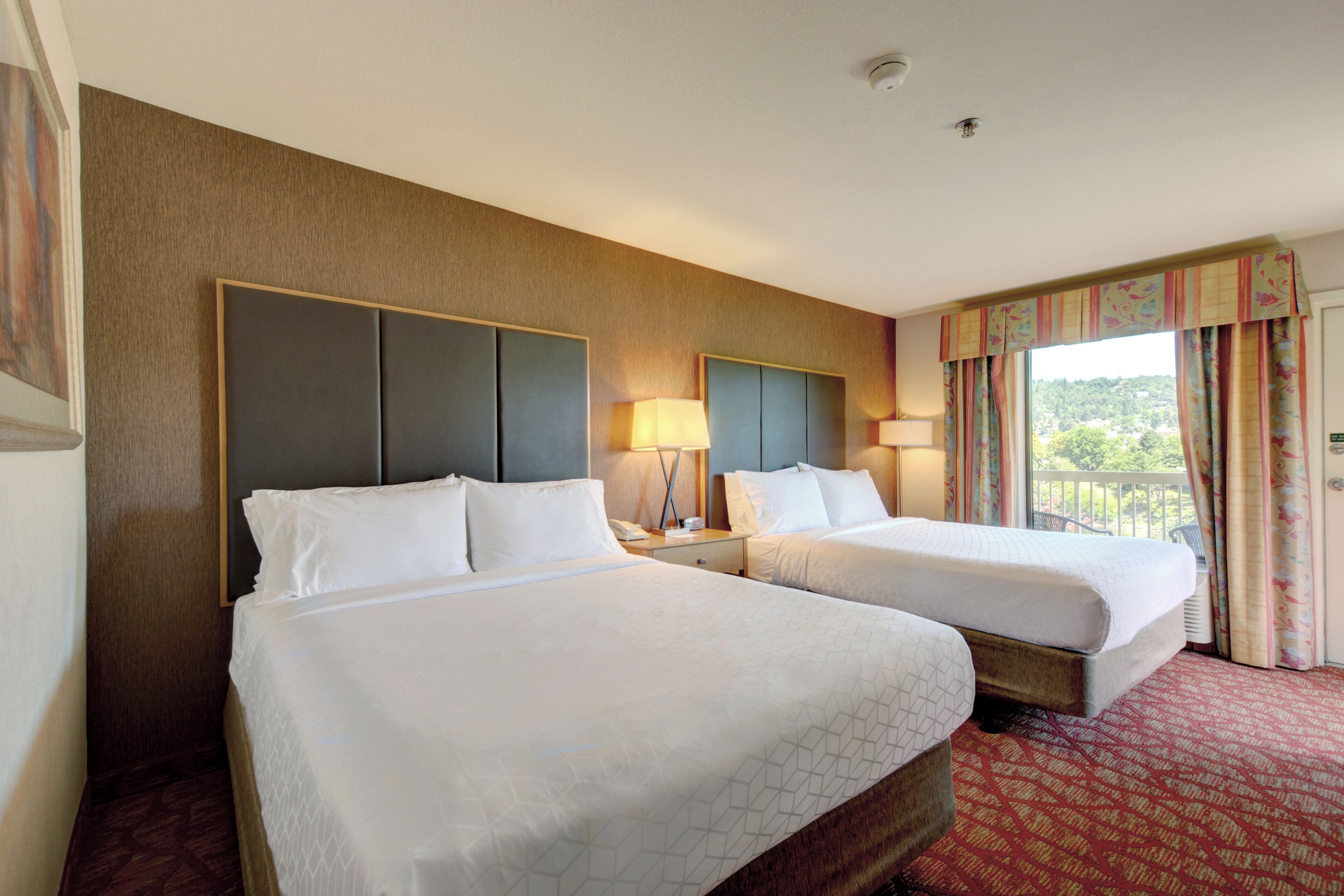 A hotel room with two beds, a large window with curtains, and a view of greenery outside.