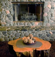 Parede de pedra com lareira e mesa de madeira com uma tigela de objetos decorativos.