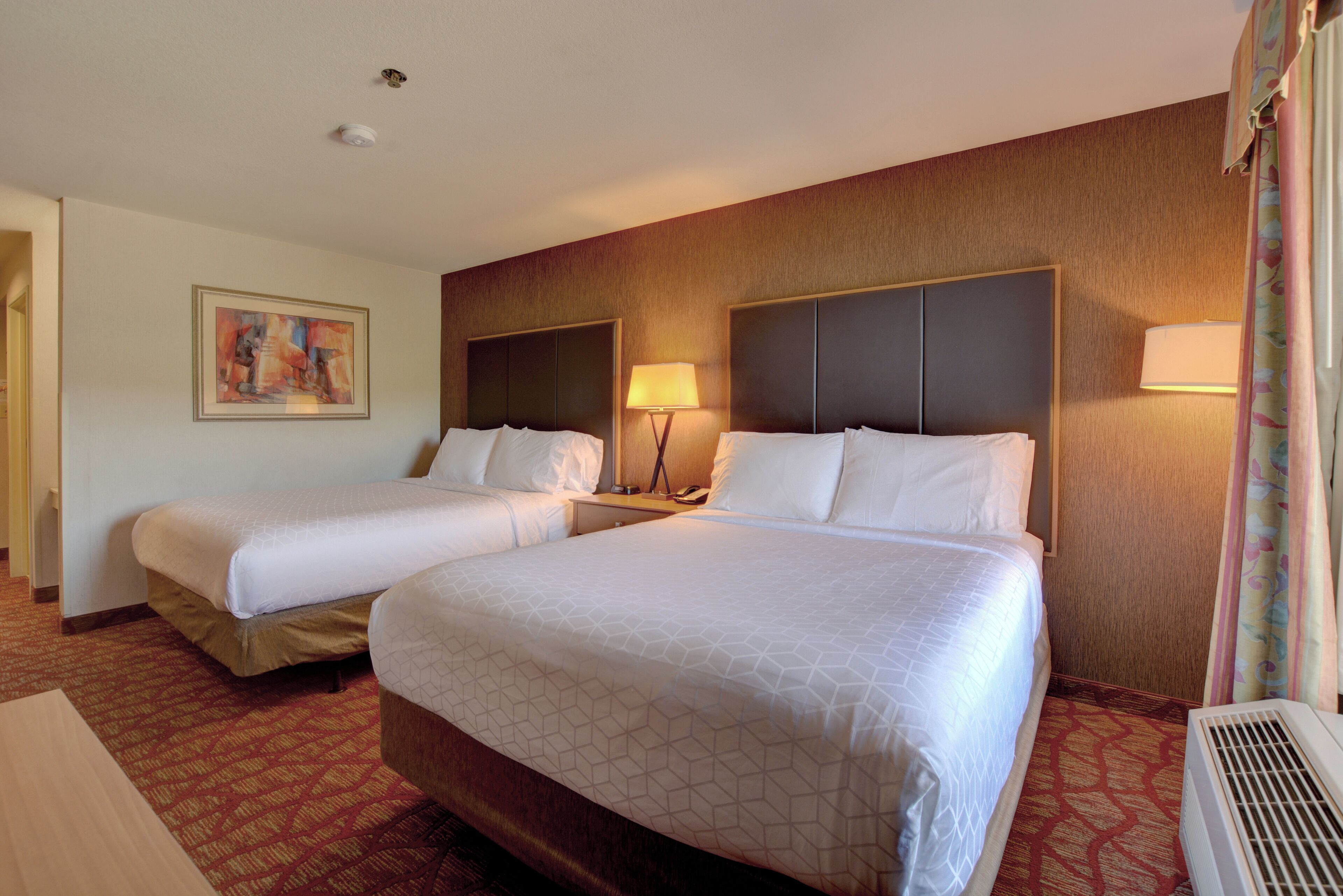 A hotel room with two beds, a painting on the wall, a lamp, and a heater.