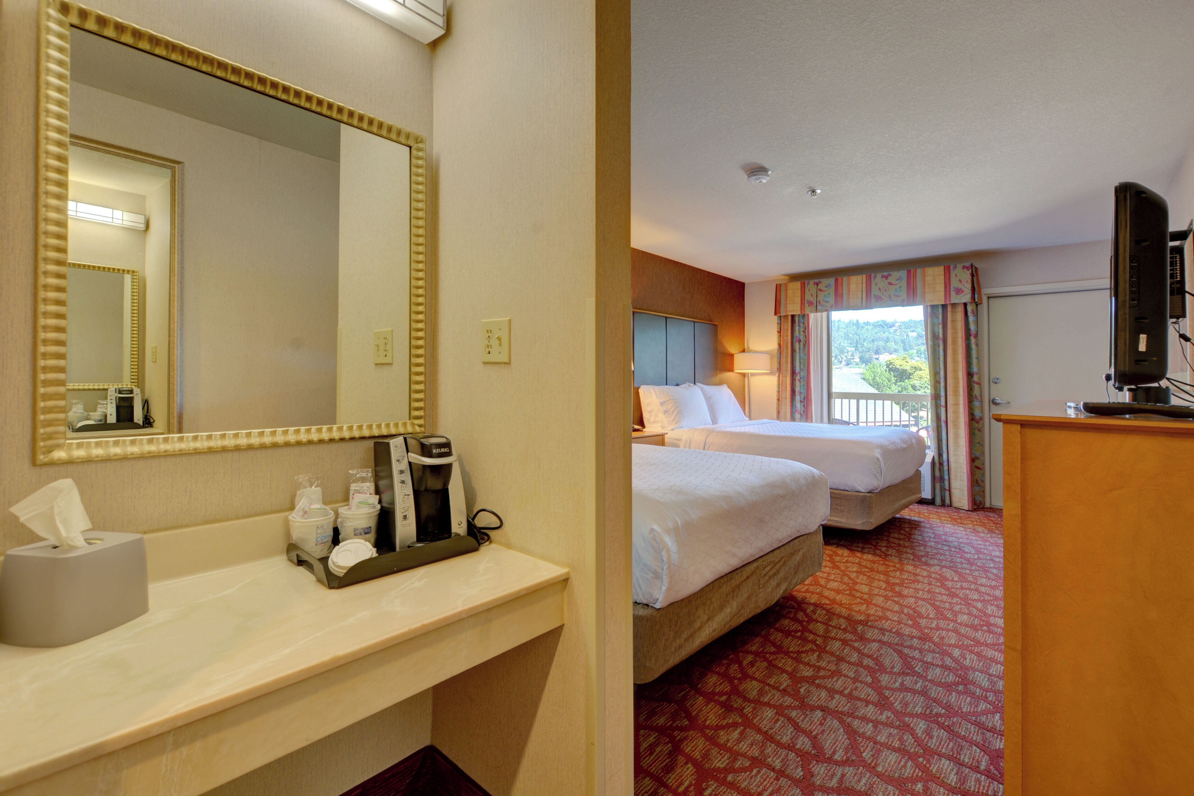 A hotel room with a large mirror, a bedside table with a coffee maker, two beds, a TV, and a window with curtains.