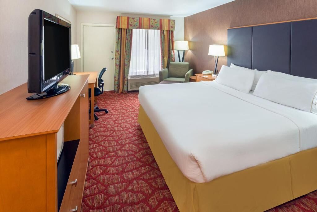 A hotel room with a bed, a desk with a television, a chair, a lamp, and a window with curtains.