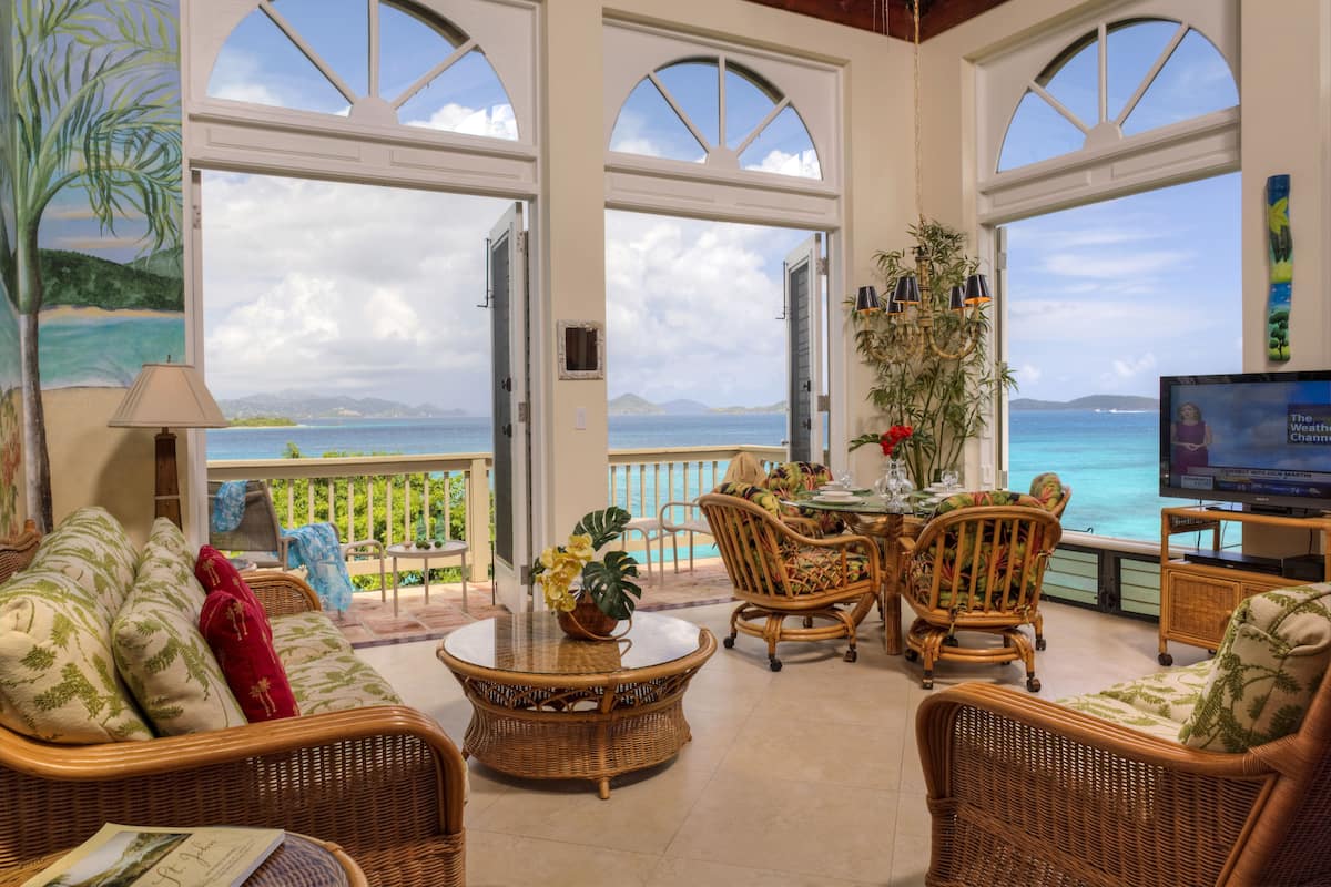 Ein geräumiges Zimmer mit großem Fenster und Blick auf einen Strand, mit Korbmöbeln, einem Fernseher und einer Topfpflanze.