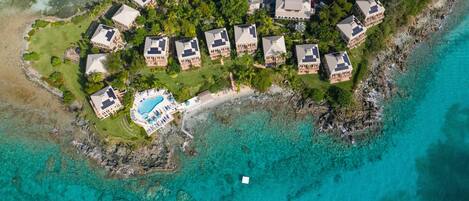 An aerial view of a coastal resort with multiple buildings, a central swimming pool, and clear turquoise waters.