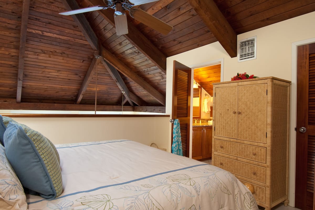 Ein Schlafzimmer mit Holzbalkendecke, einem Bett mit floralem Bettbezug, einem Korbschrank und einem Fenster mit Jalousien.