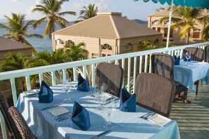 An outdoor dining area with tables set for two, overlooking a beach and palm trees.