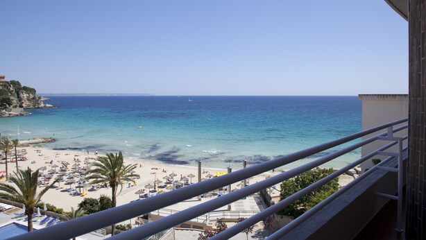 A beach view from a balcony with clear skies, a sandy beach, umbrellas, and palm trees. - Be Live Experience Costa Palma (Palma de Mallorca)