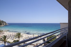 Vista de una playa desde un balcón, con cielos despejados, arena, sombrillas y palmeras.
