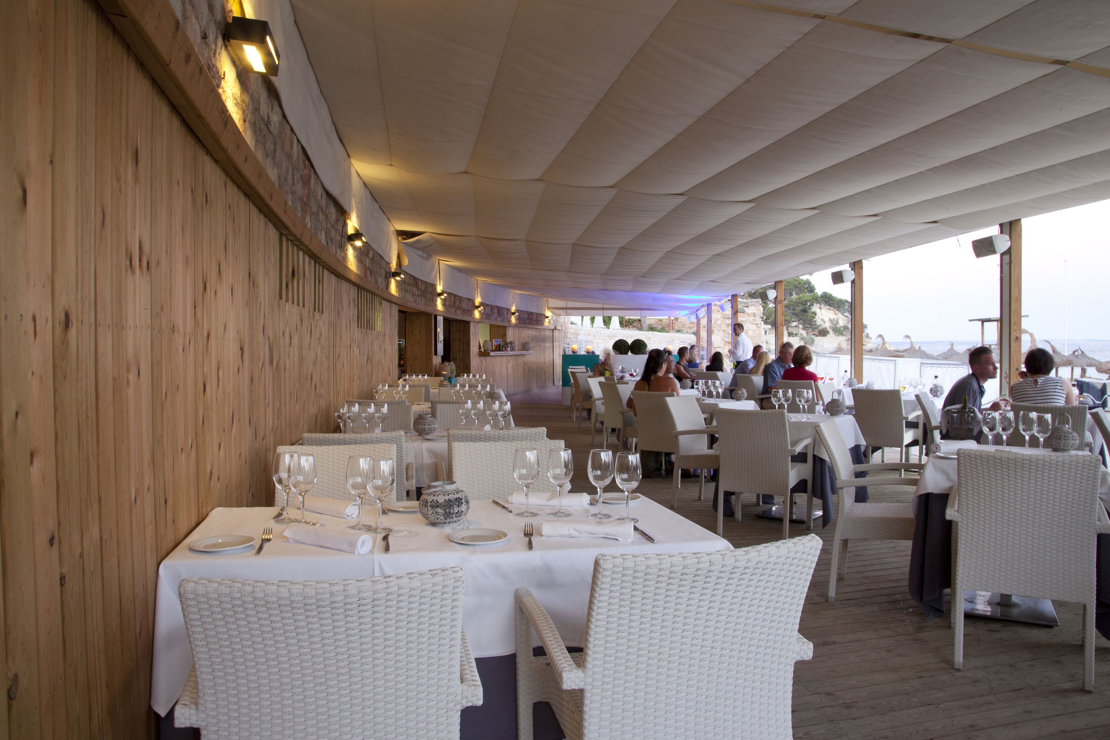 Un restaurante con varias mesas preparadas para comer, con sillas de mimbre y manteles blancos.