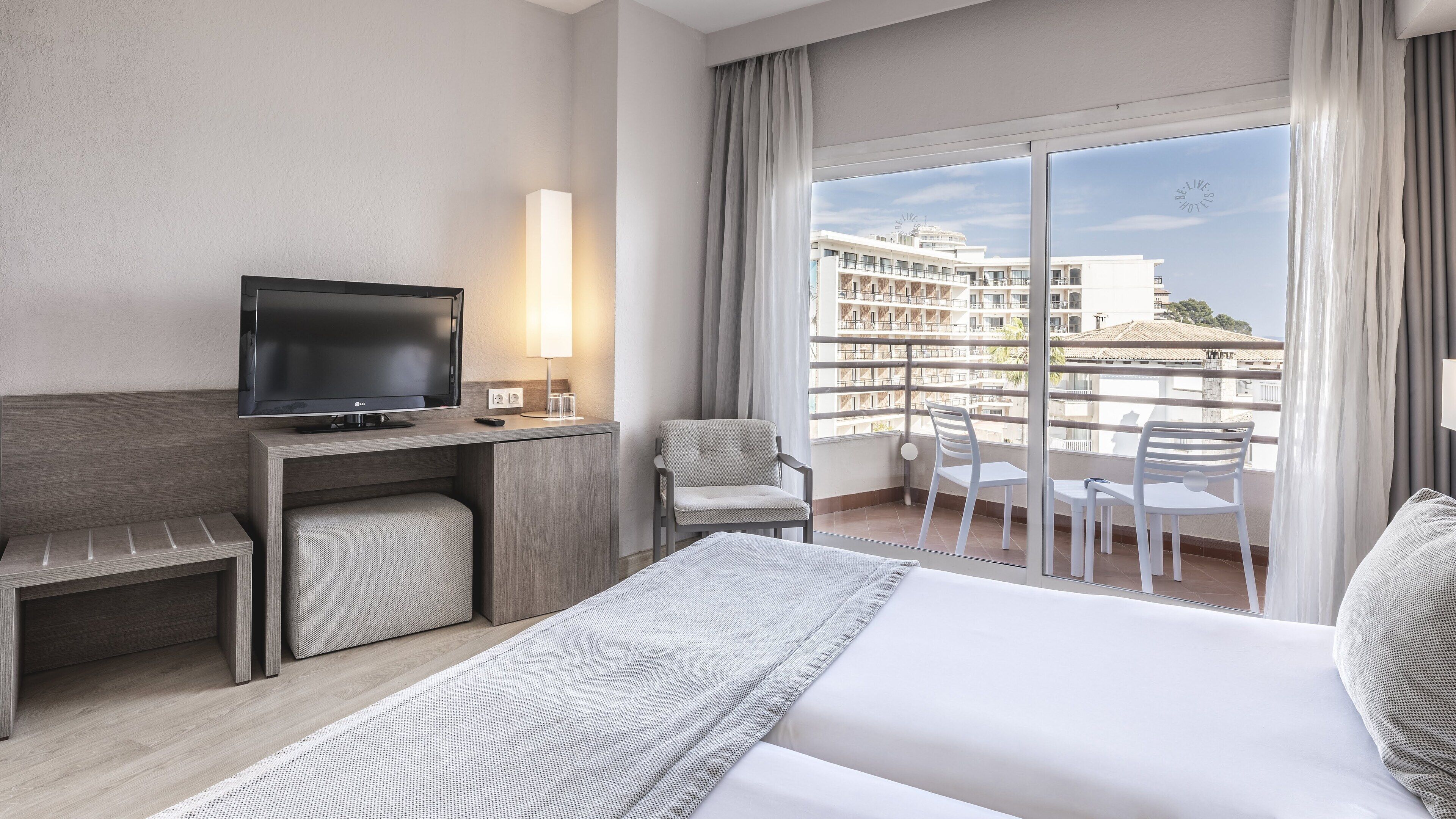 A hotel room with a bed, a desk with a TV, a chair, and a balcony with a view of buildings.