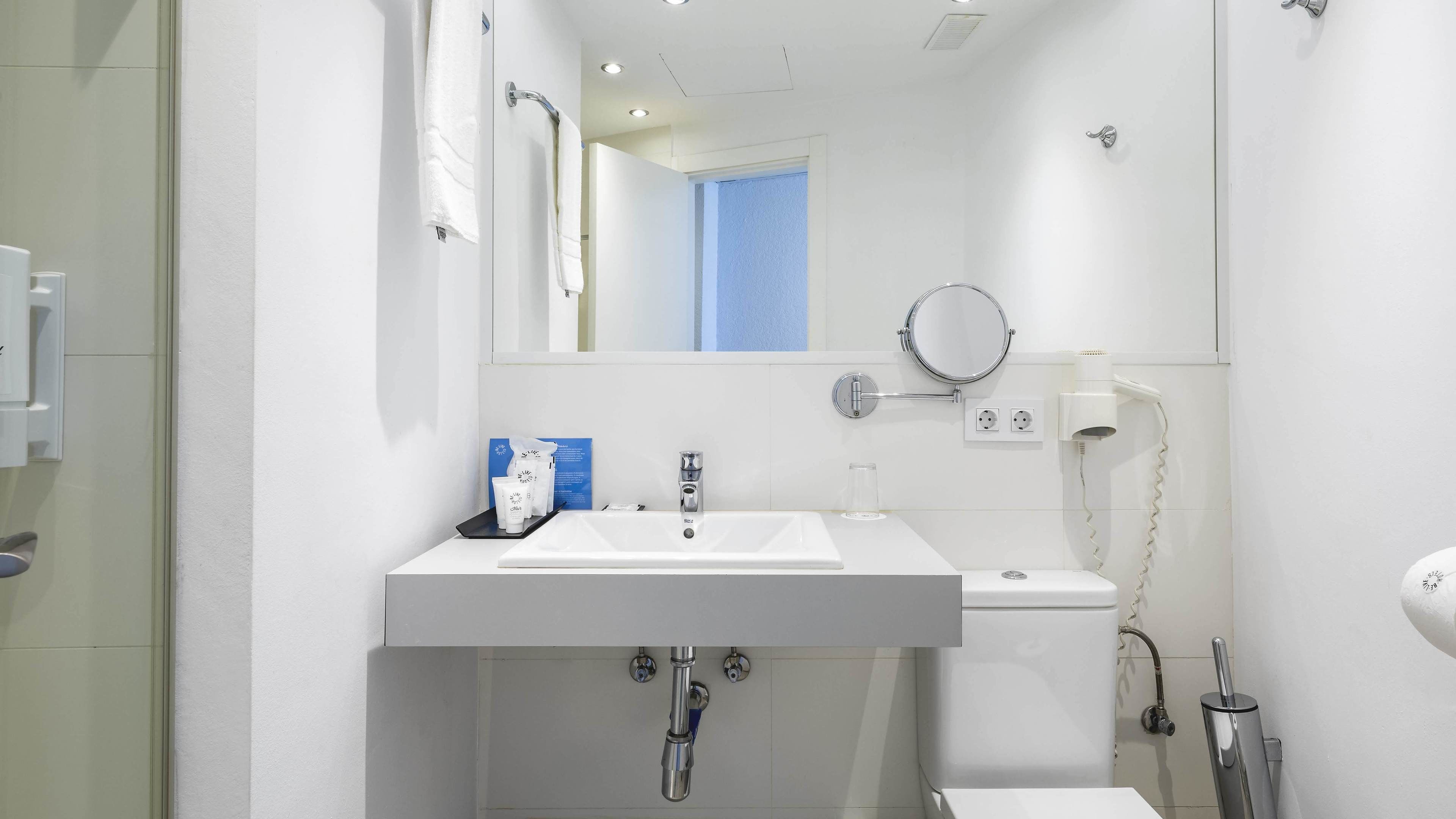 Ein modernes Badezimmer mit einem weißen Waschbecken, Spiegel und Toilette. Auf dem Waschbecken liegen Toilettenartikel, es gibt einen Handtuchhalter und einen Haartrockner an der Wand.