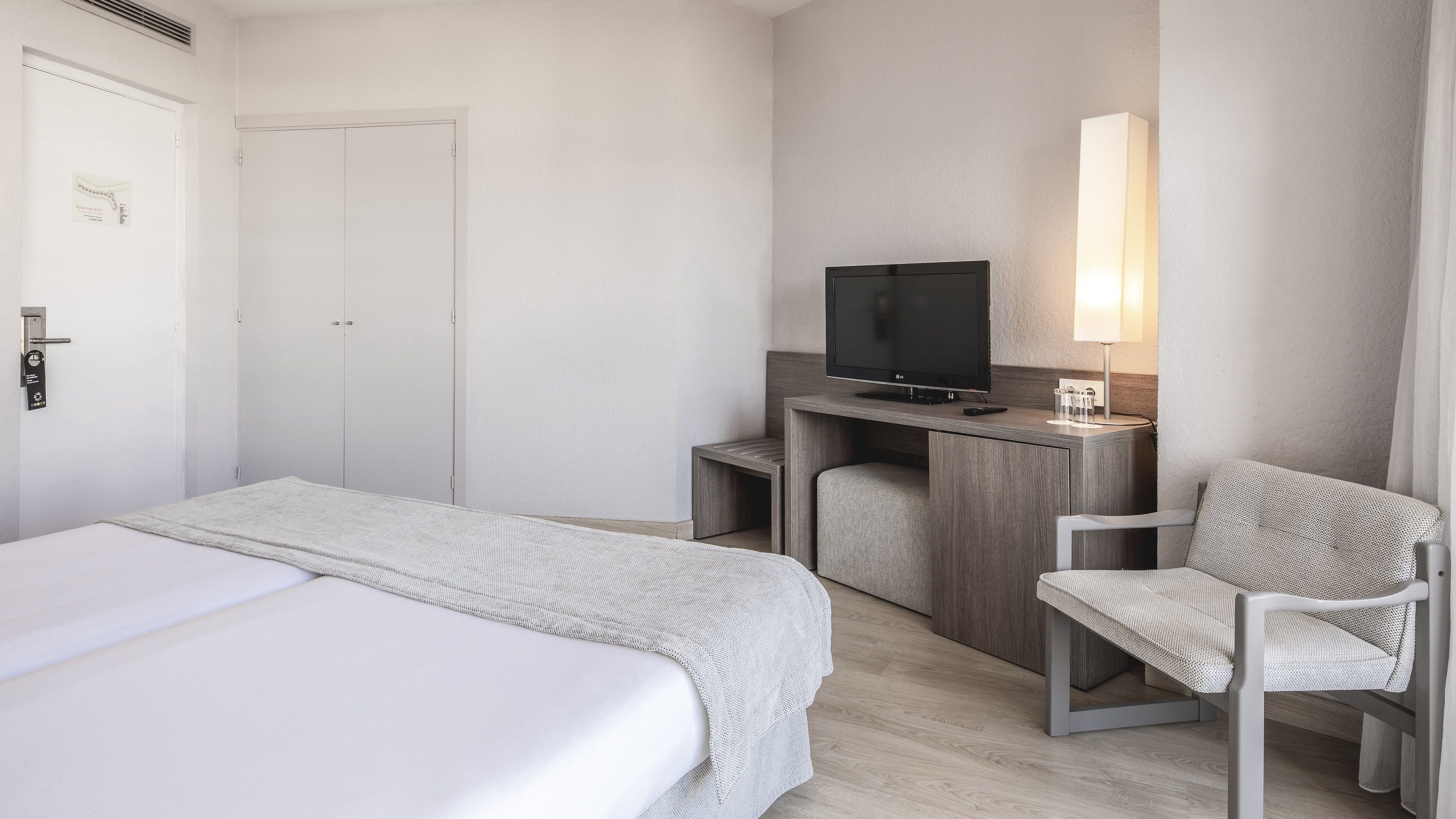 Habitación de hotel con dos camas, un escritorio con televisor, una silla y una mesita de noche con lámpara.