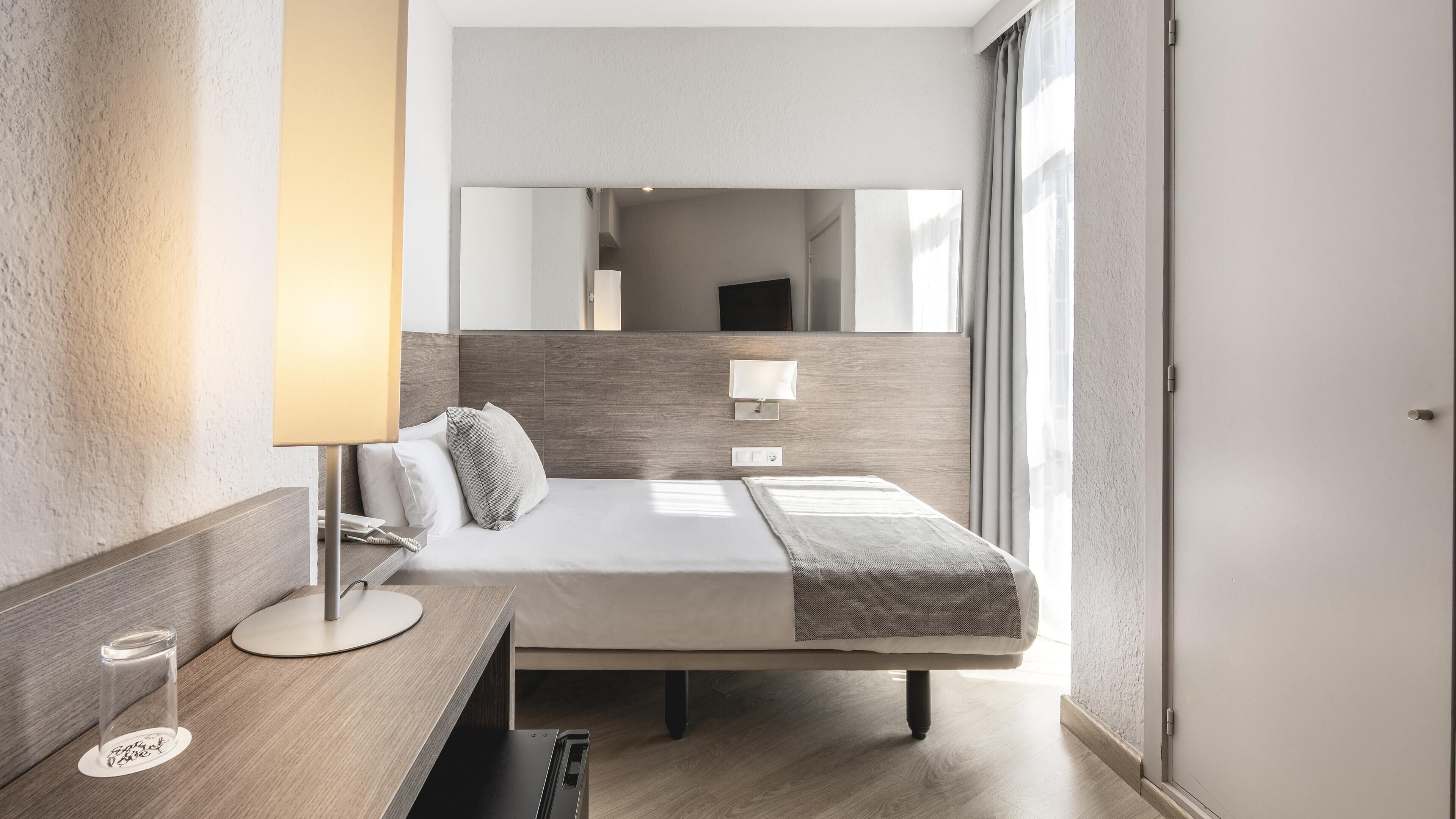 Una habitación de hotel moderna con cabecera de madera, una mesita de noche con lámpara, una cama con manta gris y un espejo grande en la pared.