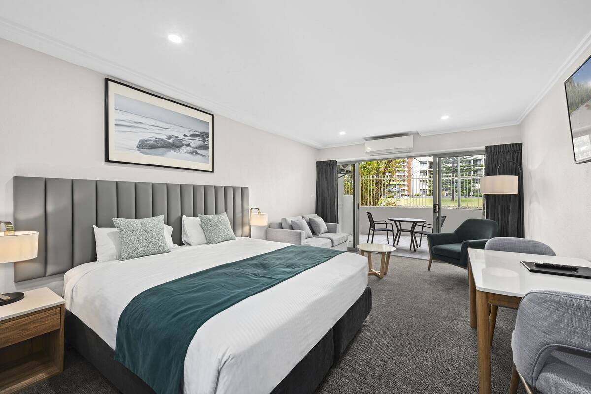 A modern hotel room with a large bed, a desk, a sofa, and a balcony with a view of the city.