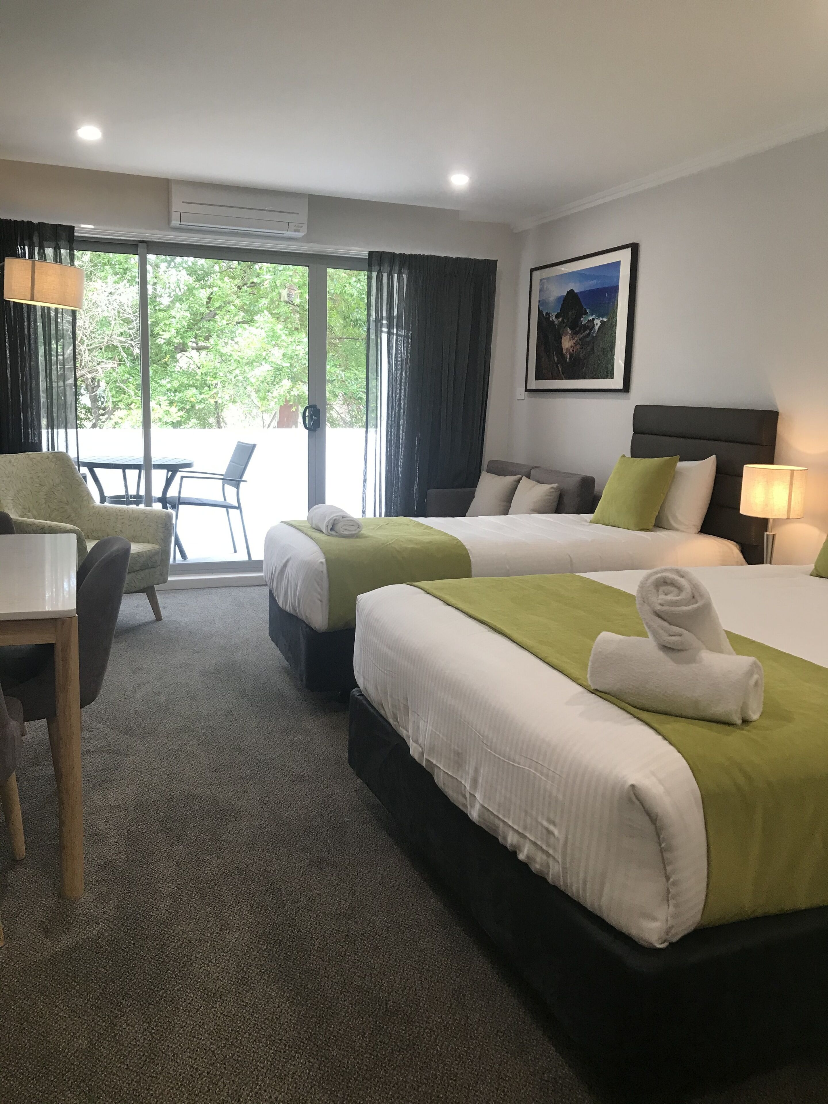 A hotel room with two beds, a desk, and a view of greenery outside.