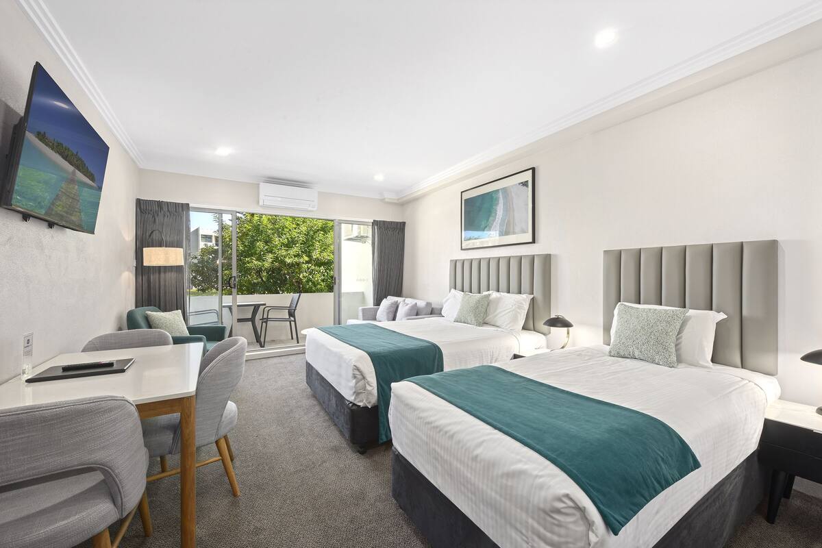 A hotel room with two beds, a desk, chairs, and a view of greenery outside.
