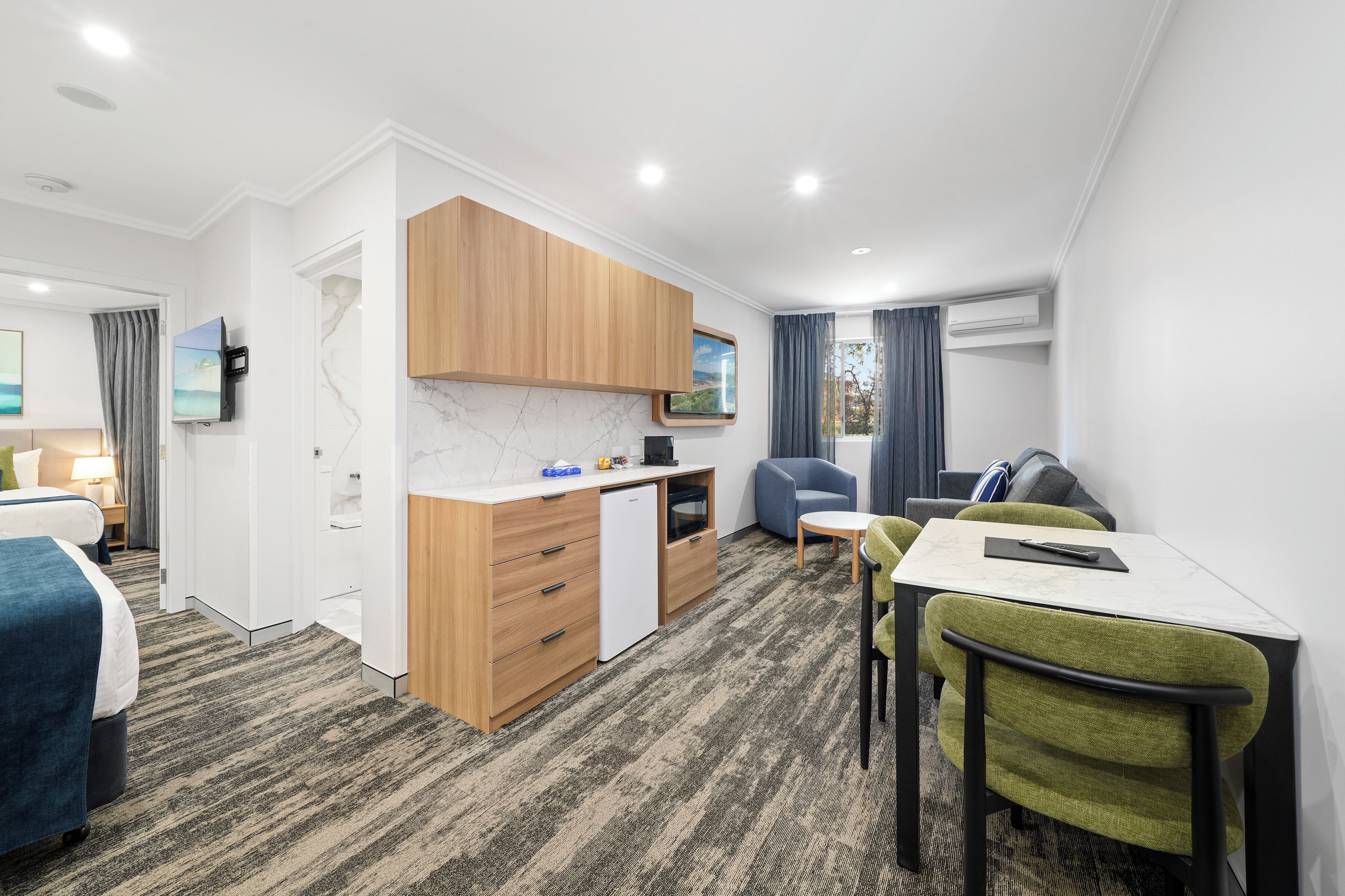 A modern hotel room with a kitchenette, a dining area, a bed, and a TV.