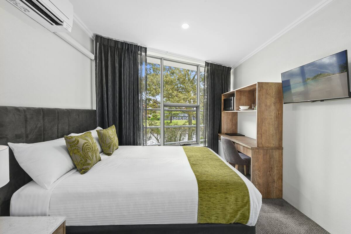 A hotel room with a large bed, a desk, a chair, a TV, and a window with curtains.
