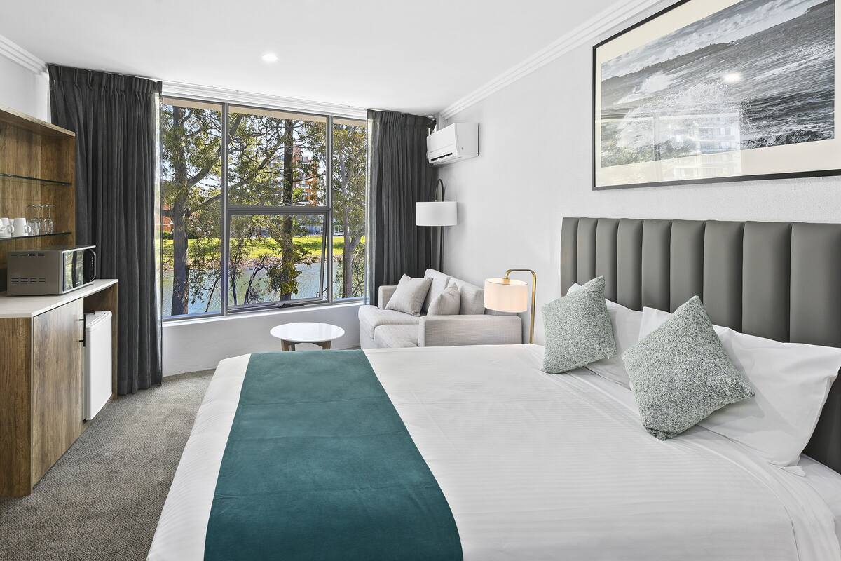 A hotel room with a large bed, a desk with a computer, a chair, a lamp, and a window with a view of trees and buildings.