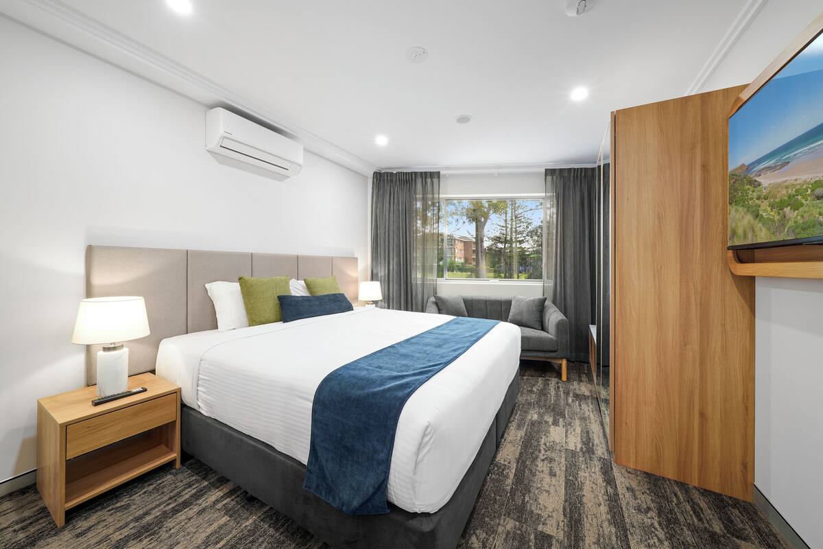 A hotel room with a bed, bedside table, lamp, and a TV on a wooden stand.