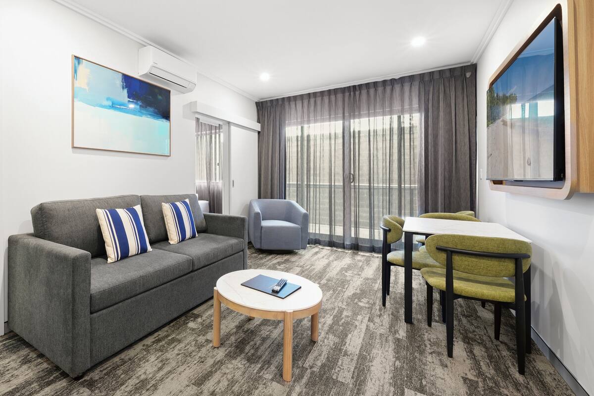 A modern living room with a grey sofa, a round coffee table, a grey armchair, a small table with a green chair, and large windows with grey curtains.