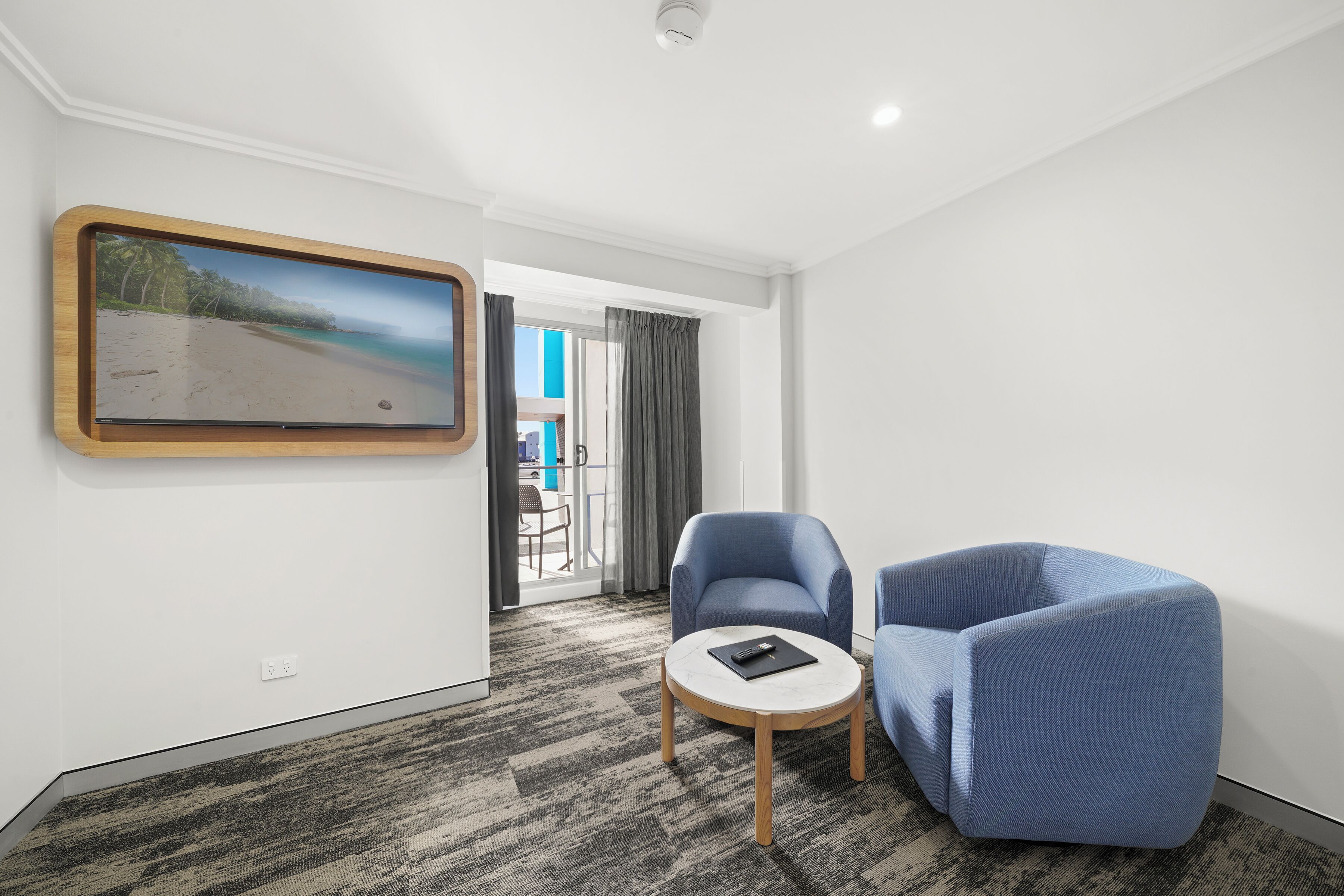 Habitación de hotel con una televisión grande de montaje en pared, dos sillones azules, una mesita redonda y una alfombra estampada.