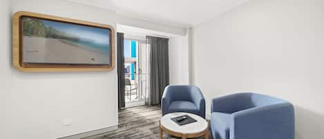 Habitación de hotel con una televisión grande de montaje en pared, dos sillones azules, una mesita redonda y una alfombra estampada.