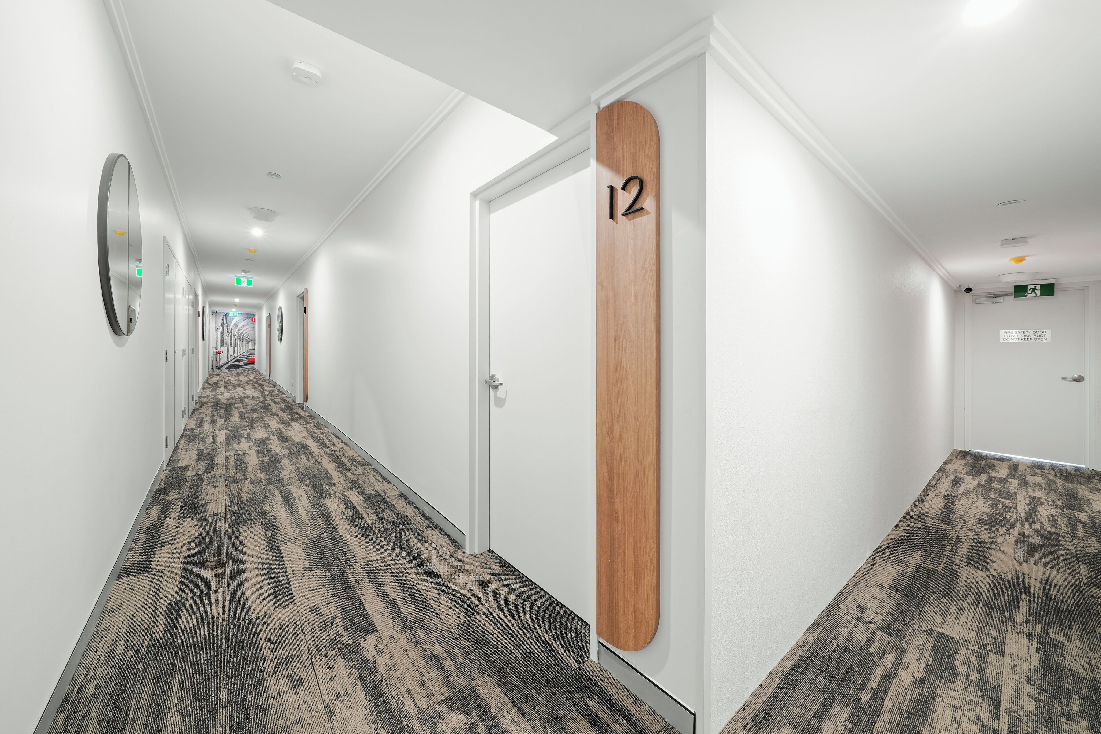 Un couloir d’hôtel avec un sol recouvert de moquette, des murs blancs et une porte en bois portant le numéro 12.