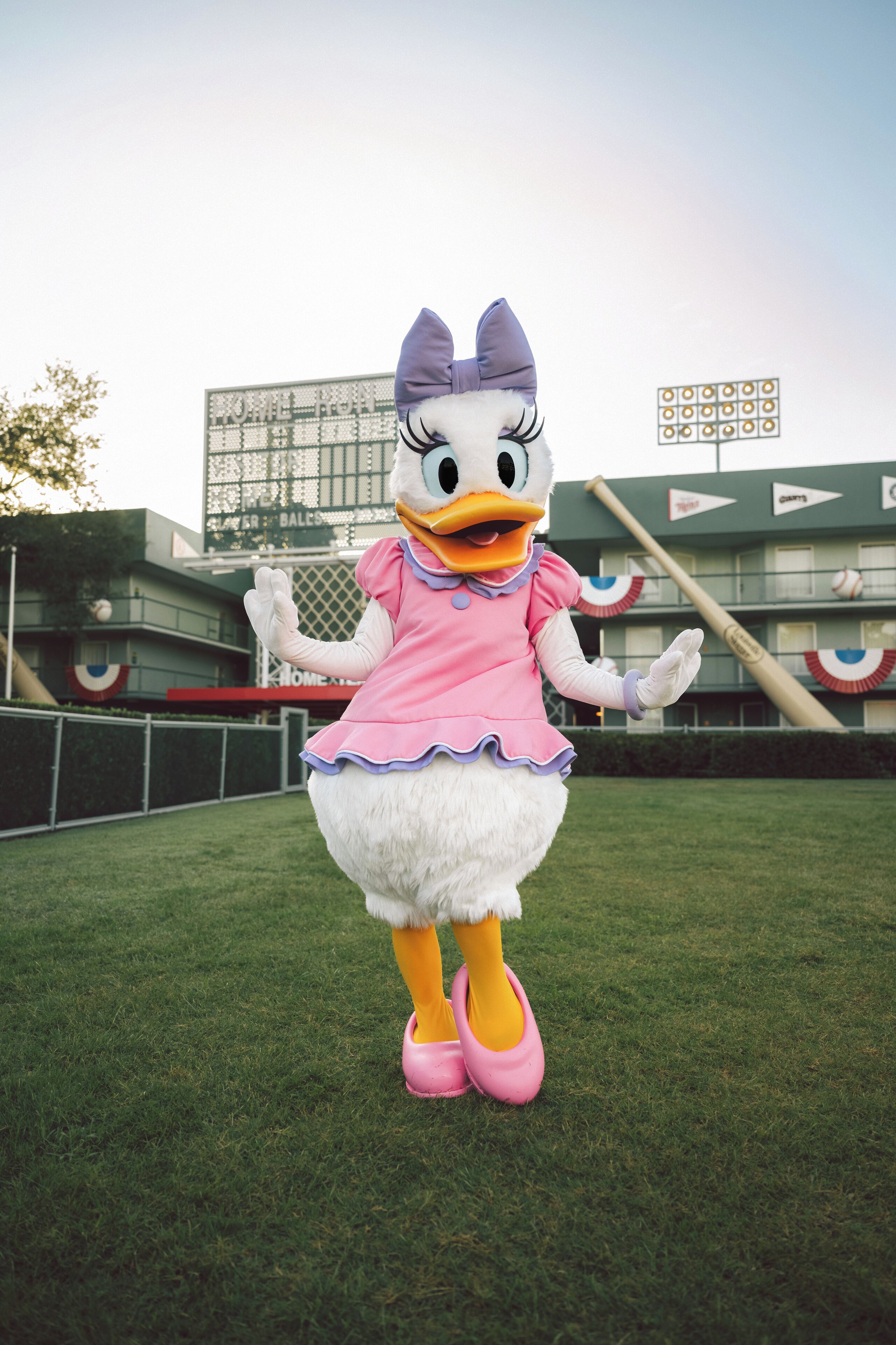A mascot in a pink dress and bow, standing on a grassy field with a baseball stadium in the background.