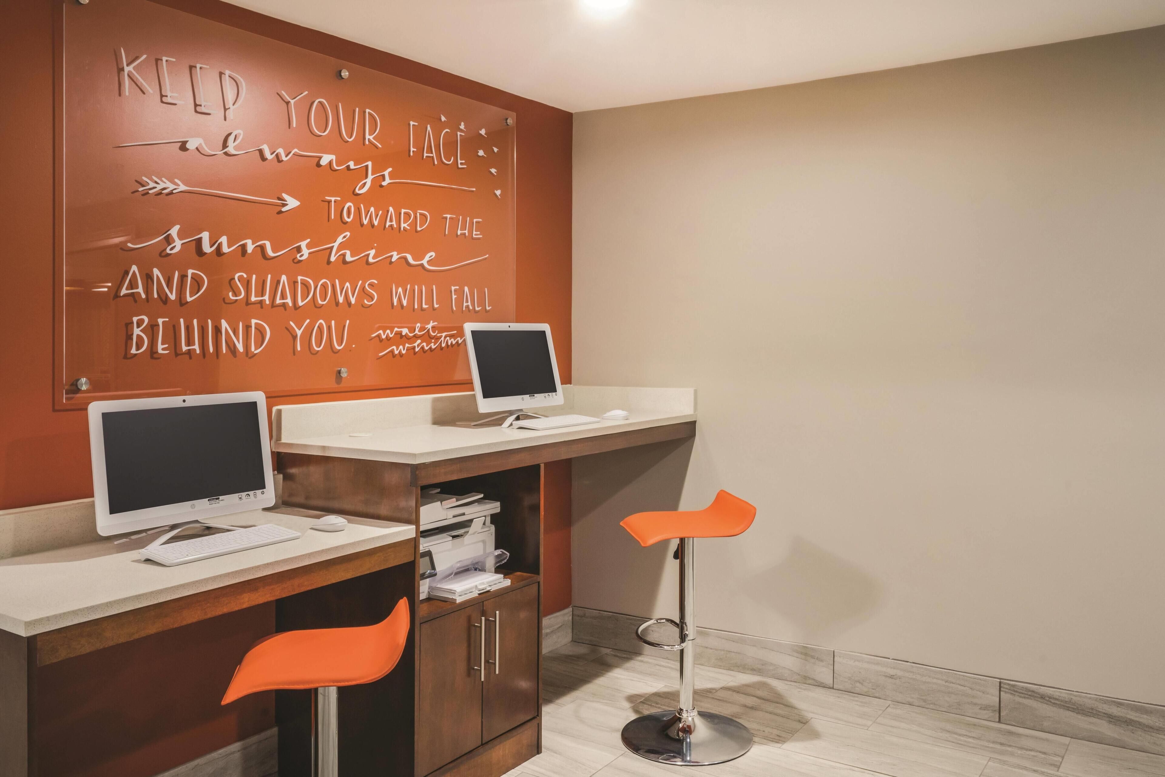 Una habitación con dos escritorios, cada uno con un ordenador y teclado, una silla, y una frase motivacional en la pared.