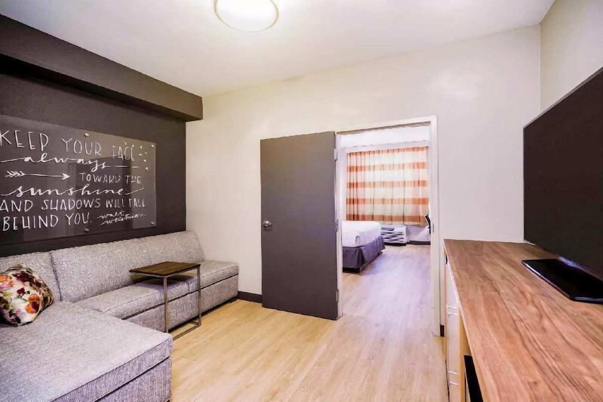 Una habitación moderna con sofá, mesa de madera, cama con cortinas a rayas y una pizarra con mensajes motivacionales.