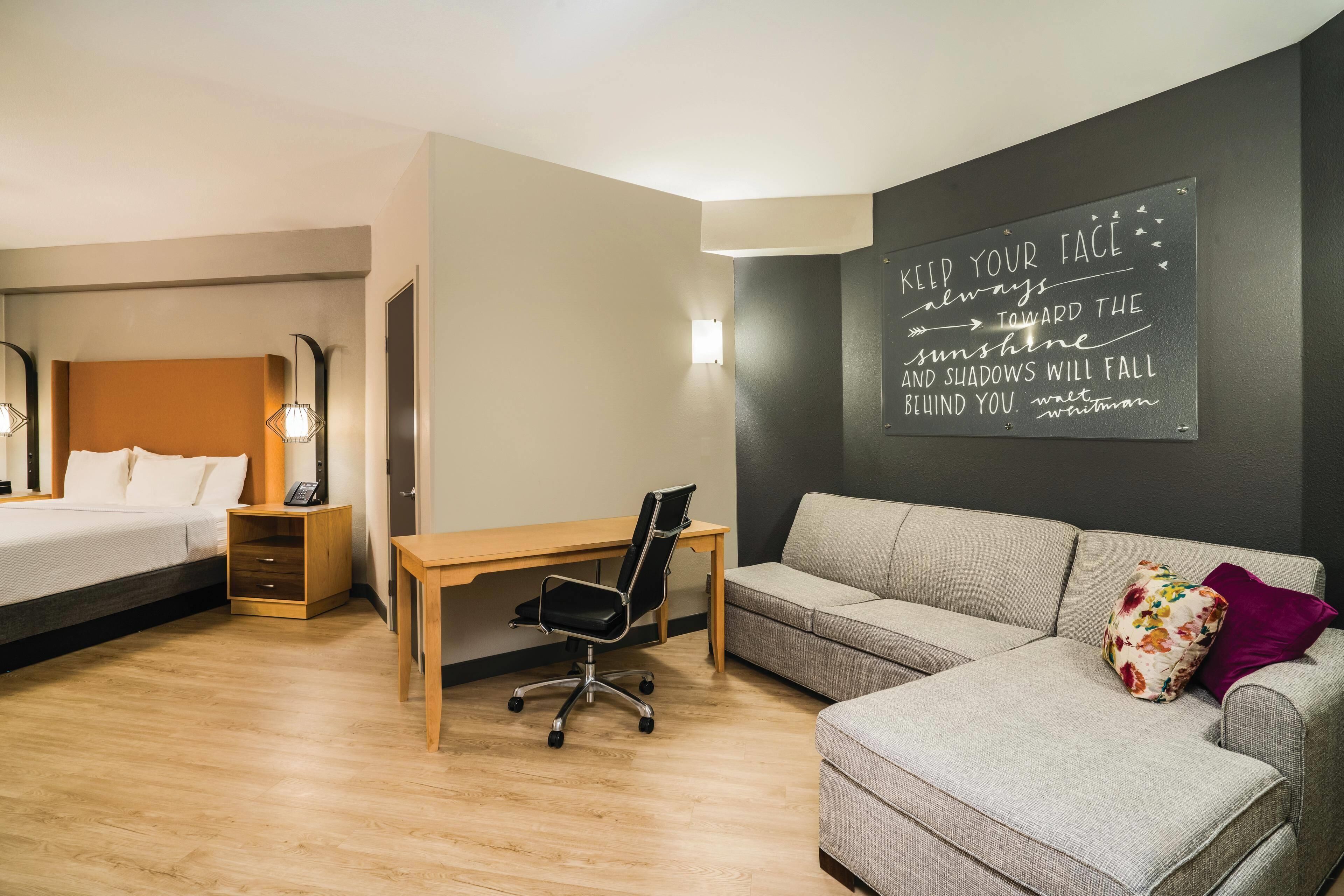 A hotel room with a bed, a desk, a chair, a sofa, and a motivational quote on the wall.
